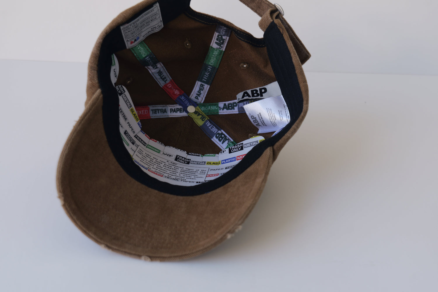 Interior view of the bABP B-SIDE brown washed cotton hat showing a removable branded cardboard insert for shape, three woven labels (including a hidden "easter egg" patch and "Speak Up Fashionable" tag), and "TRASH/METAL/PLASTIC" branded seam tapes