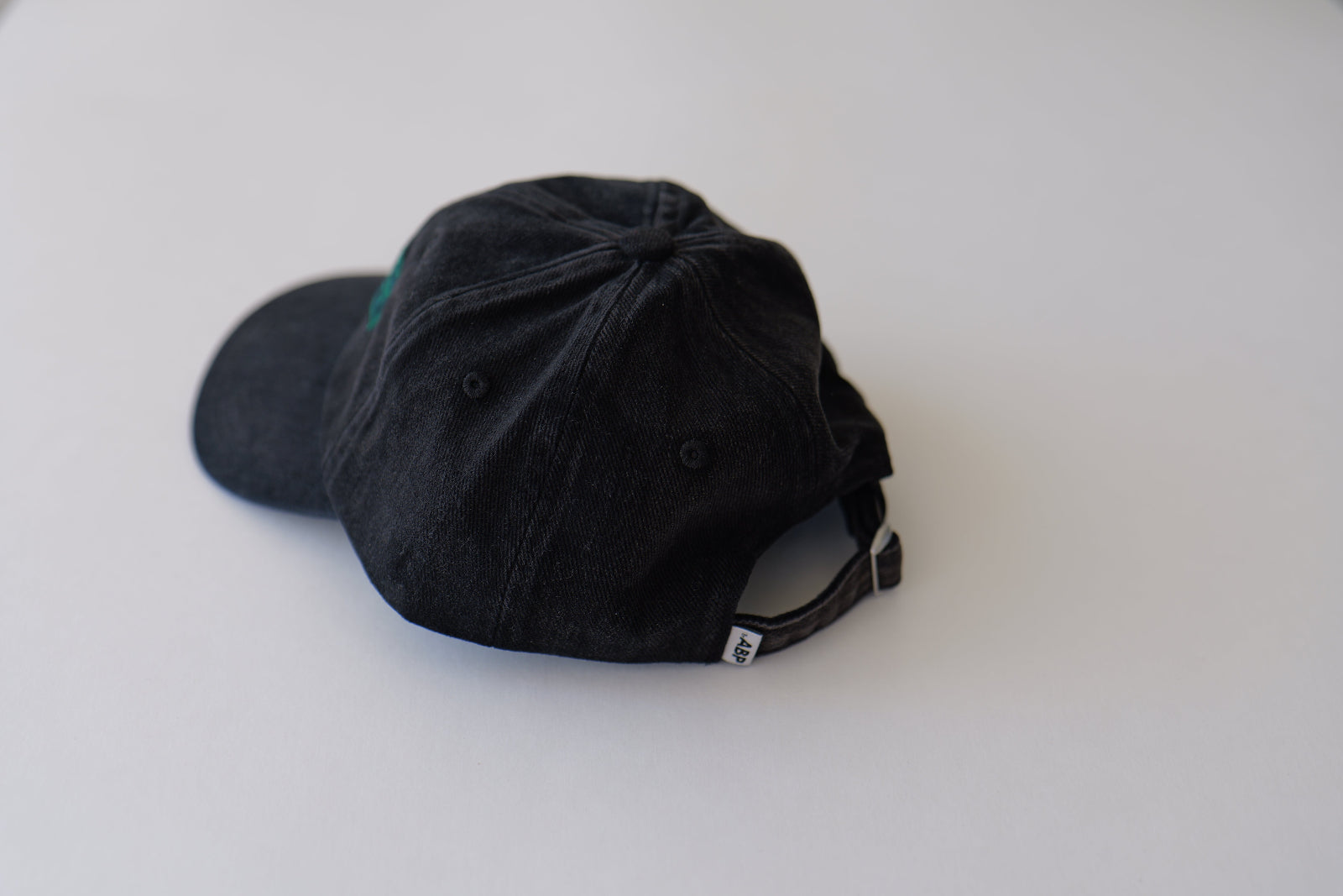 Rear angled studio view of the bABP B-SIDE "COMPOST" black washed cotton hat on a light gray background, showing the adjustable back strap and woven logo tag detail