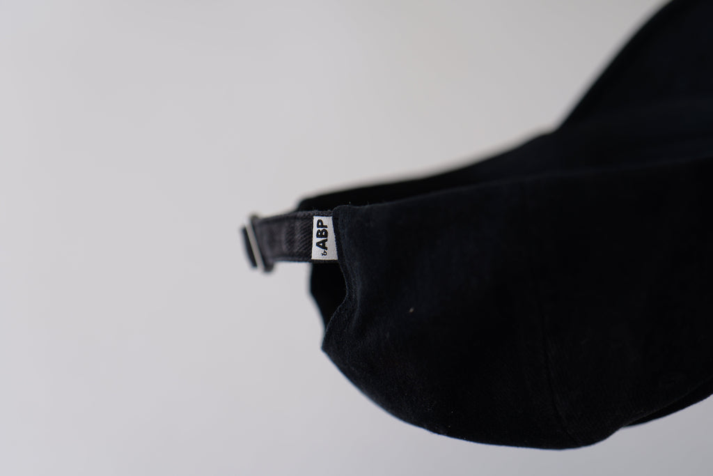 Close-up of the adjustable back strap and metal buckle on the bABP B-SIDE "COMPOST" black washed cotton hat featuring the signature white woven logo tag