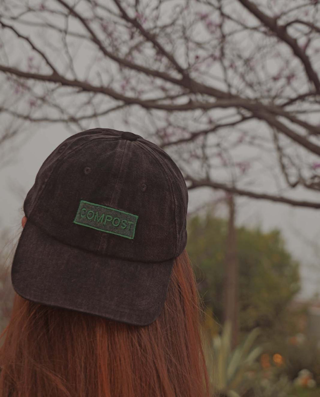 Back view of a person with long hair wearing the bABP B-SIDE "COMPOST" black washed cotton hat with a green logo patch against a background of bare tree branches