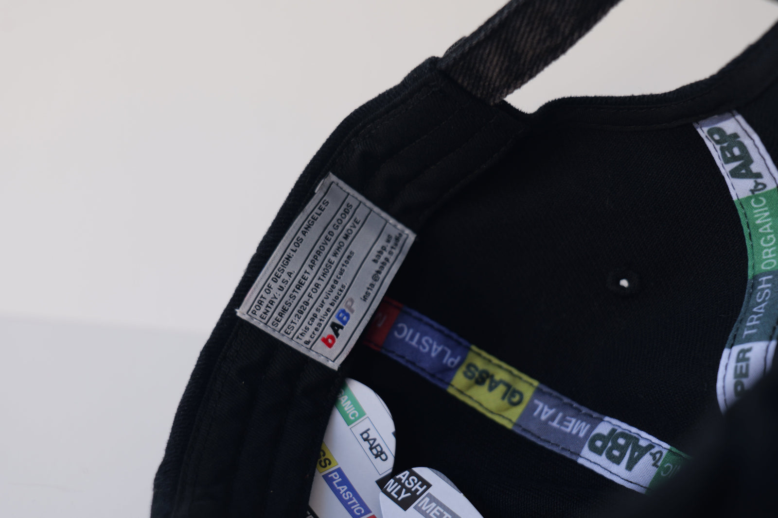 Close-up interior view of the bABP B-SIDE "COMPOST" black hat with a green logo, showing branded "TRASH/METAL/PLASTIC" seam tapes and a hidden "easter egg" woven label with street-approved goods information