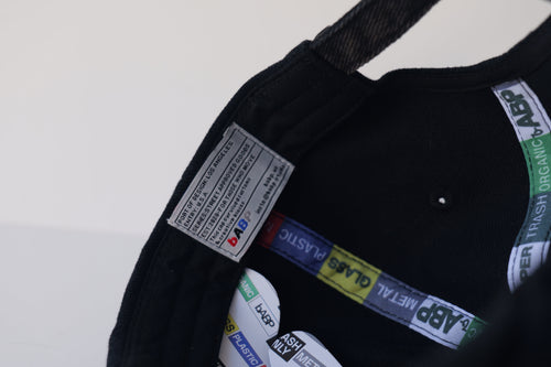 Close-up interior view of the bABP B-SIDE "COMPOST" black hat with a green logo, showing branded "TRASH/METAL/PLASTIC" seam tapes and a hidden "easter egg" woven label with street-approved goods information