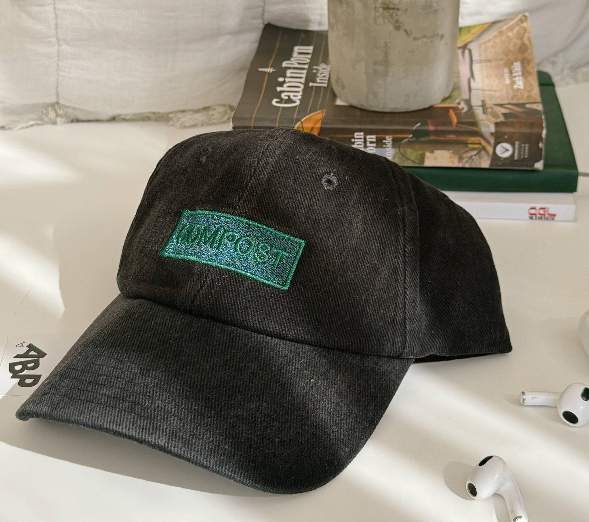 Angled view of bABP "COMPOST" black washed cotton dad hat with green embroidery from the B-SIDE collection, styled with wireless earbuds and a book on a white surface with plant shadows