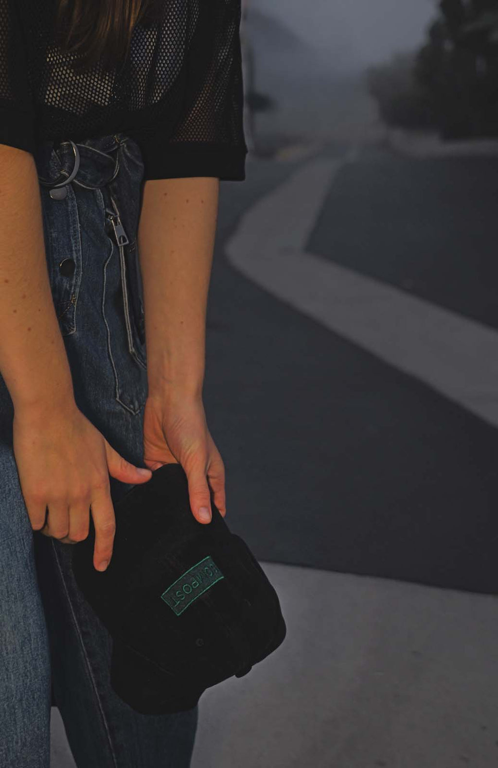 Model in a denim jumpsuit holding the bABP B-SIDE "COMPOST" black washed cotton hat with a green logo patch against a moody foggy road background