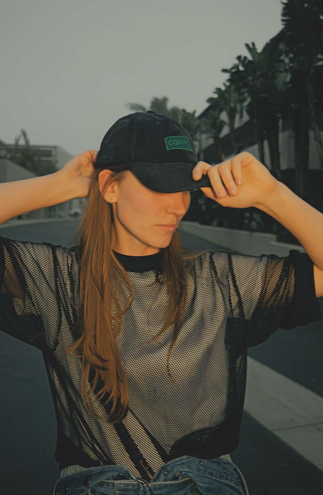 Model in a black mesh top and denim jeans posing with arms up wearing the bABP B-SIDE "COMPOST" black washed cotton hat with a green logo patch in an urban outdoor setting