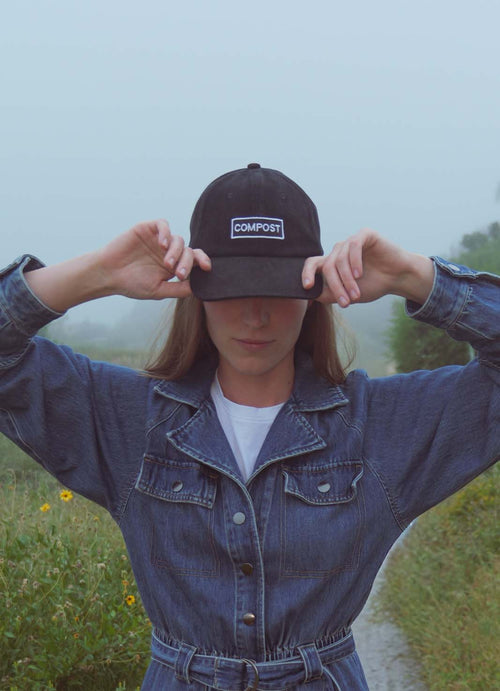 A person wearing a bABP B-SIDE "COMPOST" black washed cotton hat with a high-contrast white logo patch and a denim jacket, standing in a field with yellow flowers under a foggy sky
