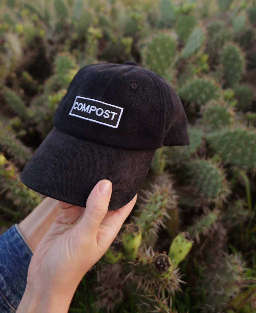 A hand holding the bABP B-SIDE "COMPOST" black washed cotton hat with a white logo patch in front of a green prickly pear cactus