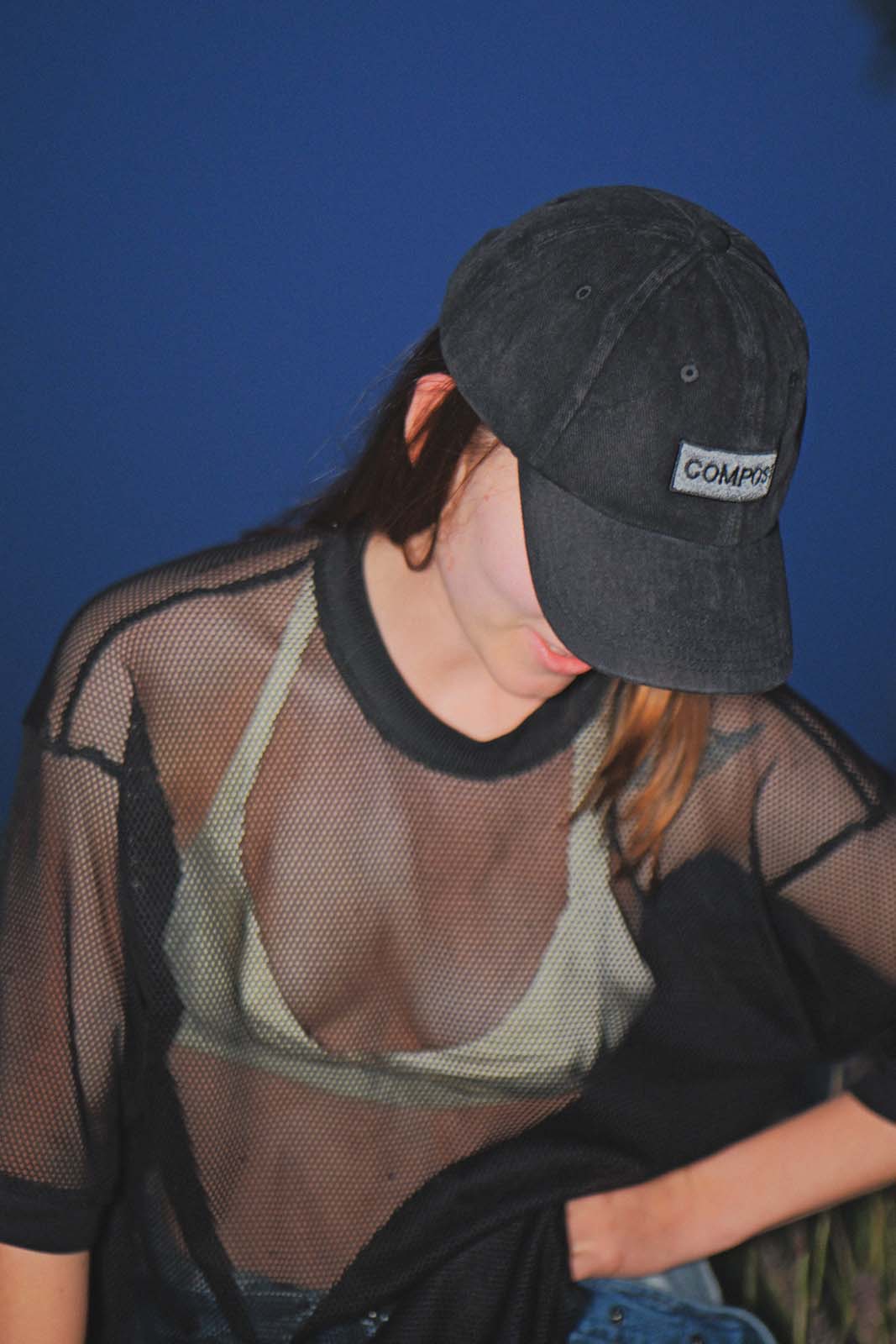 Lifestyle portrait of a person wearing the bABP B-SIDE black washed cotton hat with a "COMPOST" patch, posing against a blue background with flash photography