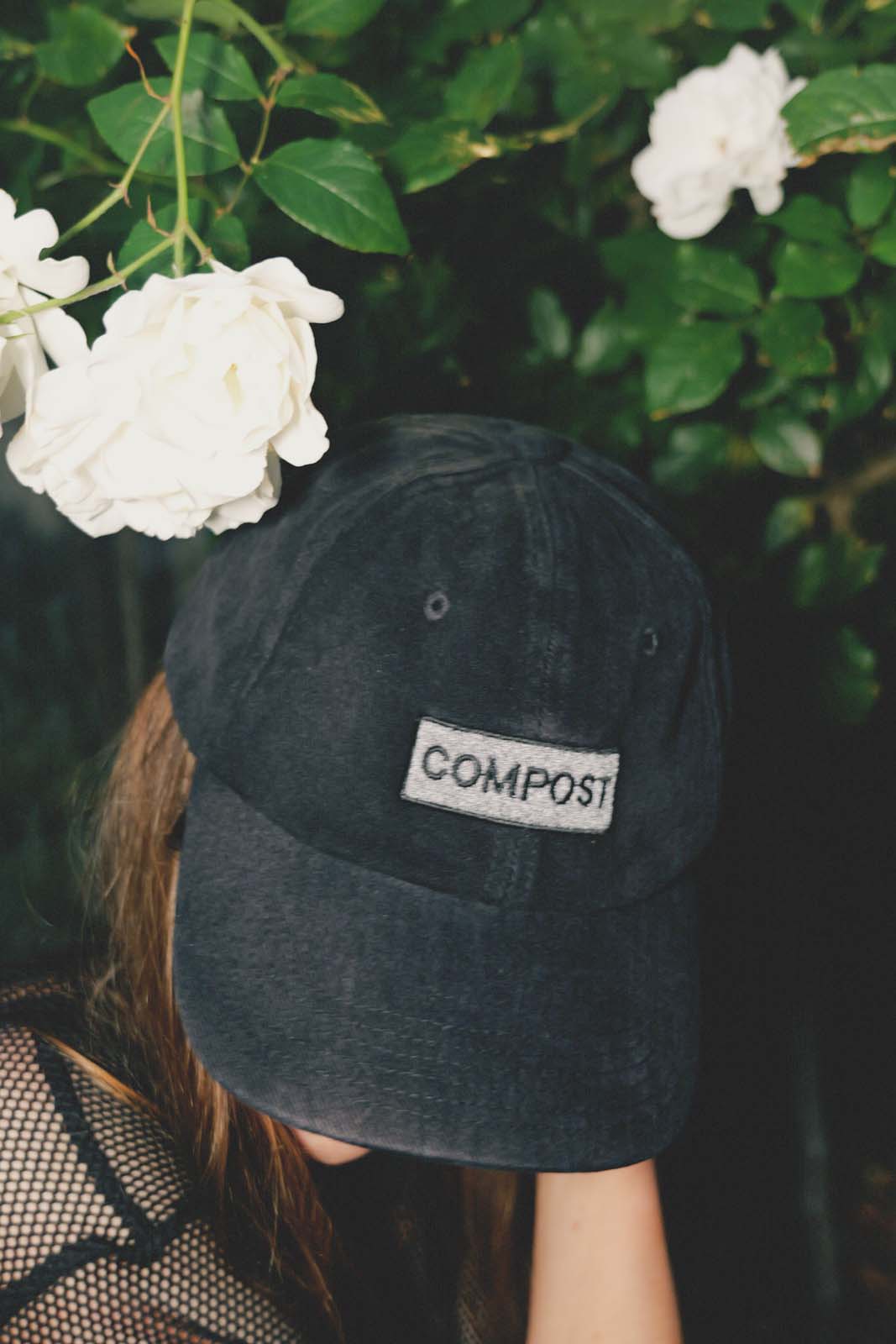 Close-up lifestyle portrait of a person wearing the bABP B-SIDE black washed cotton hat with a "COMPOST" patch, featuring white roses in the background