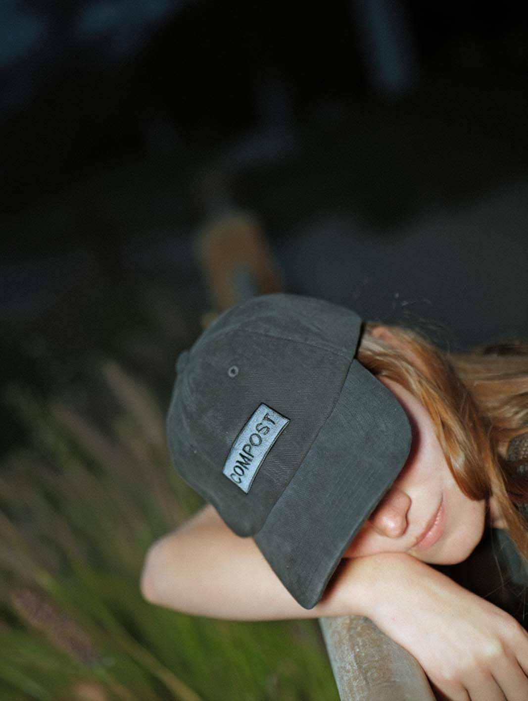 Close-up lifestyle portrait of a person wearing the bABP B-SIDE black washed cotton hat with a gray "COMPOST" patch, leaning on a wooden rail outdoors