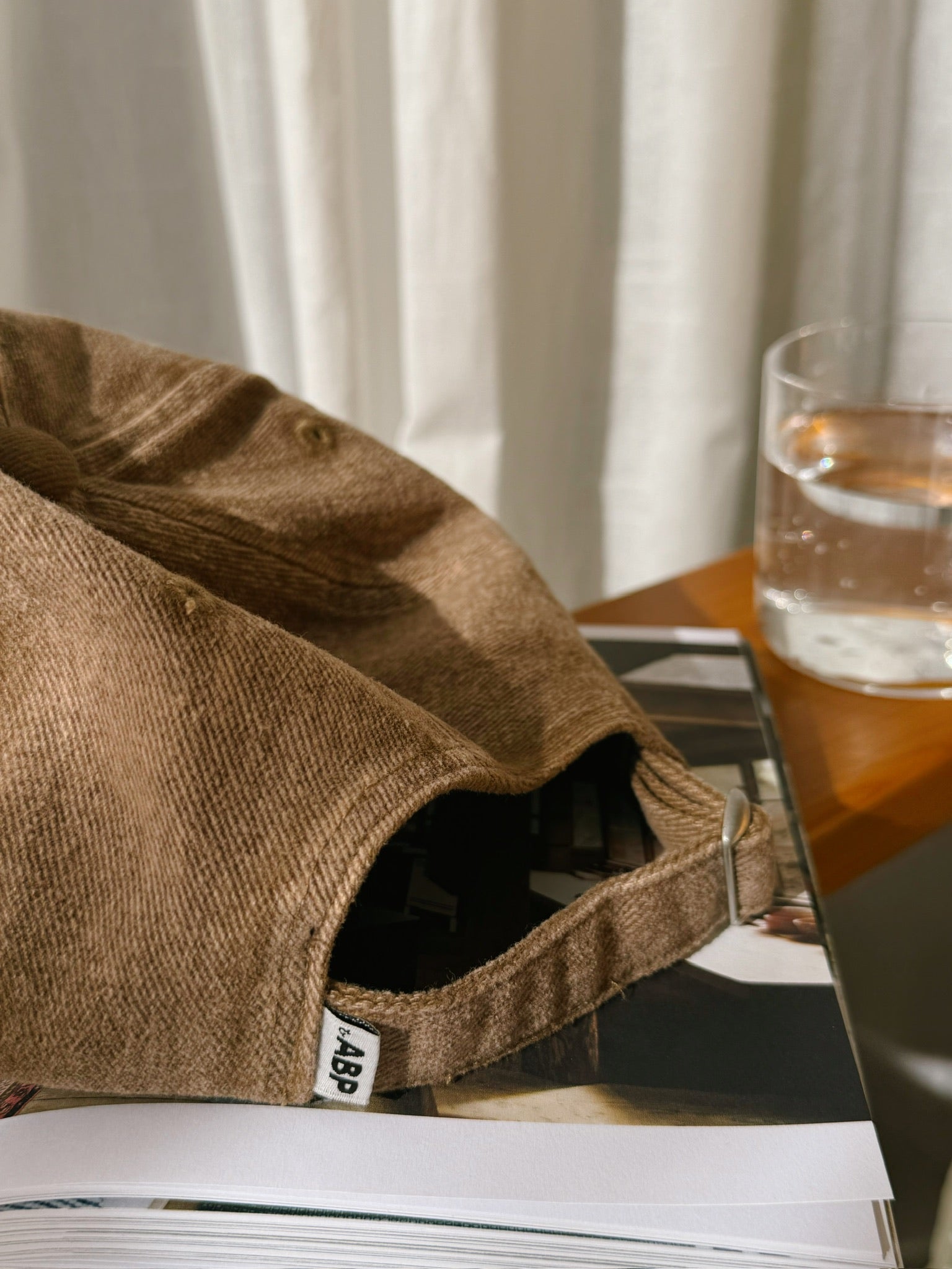 Close-up of bABP "GARBAGE" tan distressed cotton dad hat from the B-SIDE collection, focusing on the branded side label and metal strap closure, styled on a magazine with a glass of water and soft window light