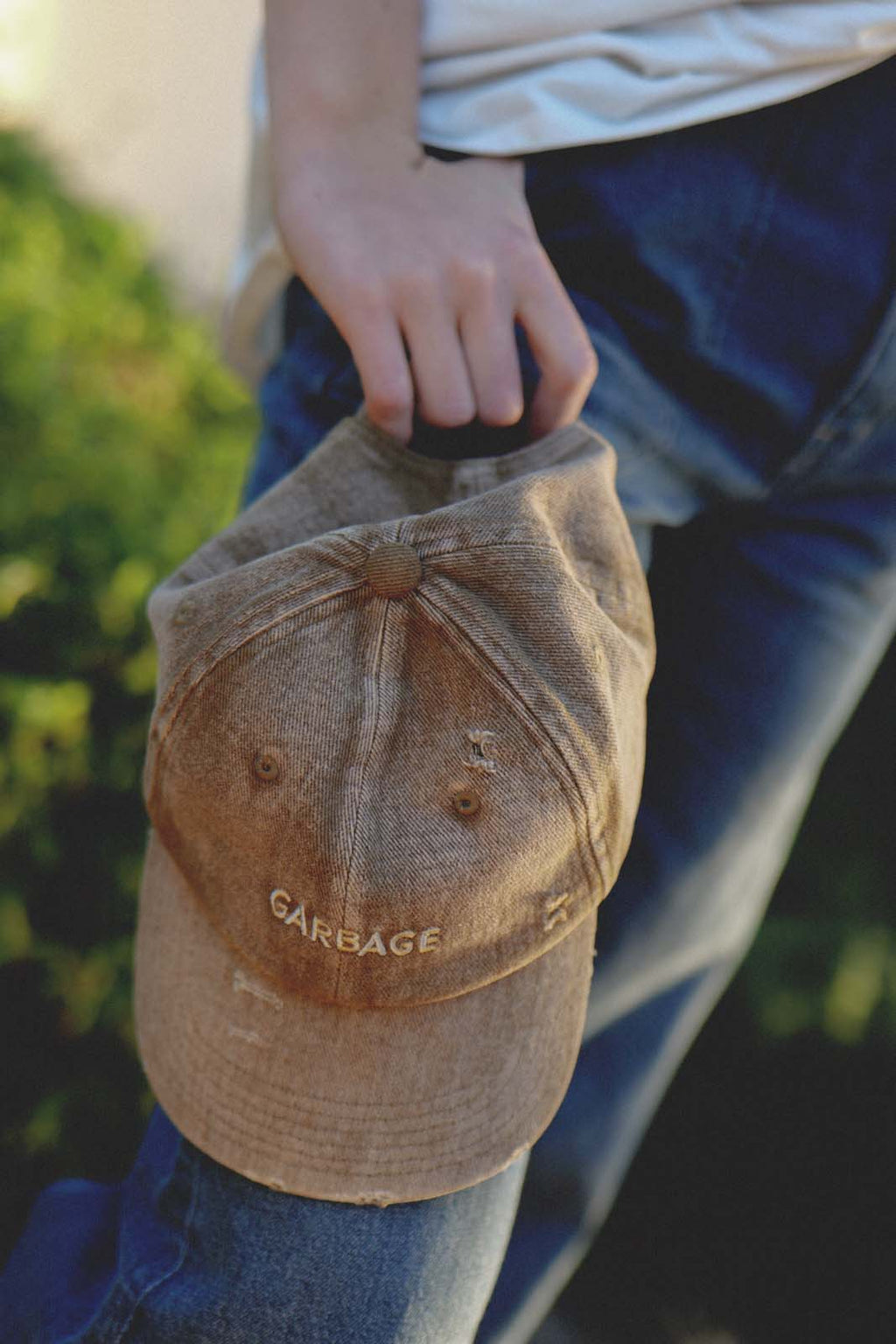 Close-up lifestyle shot of a person's hand holding the bABP B-SIDE tan washed cotton hat with "GARBAGE" embroidery against a blue denim background