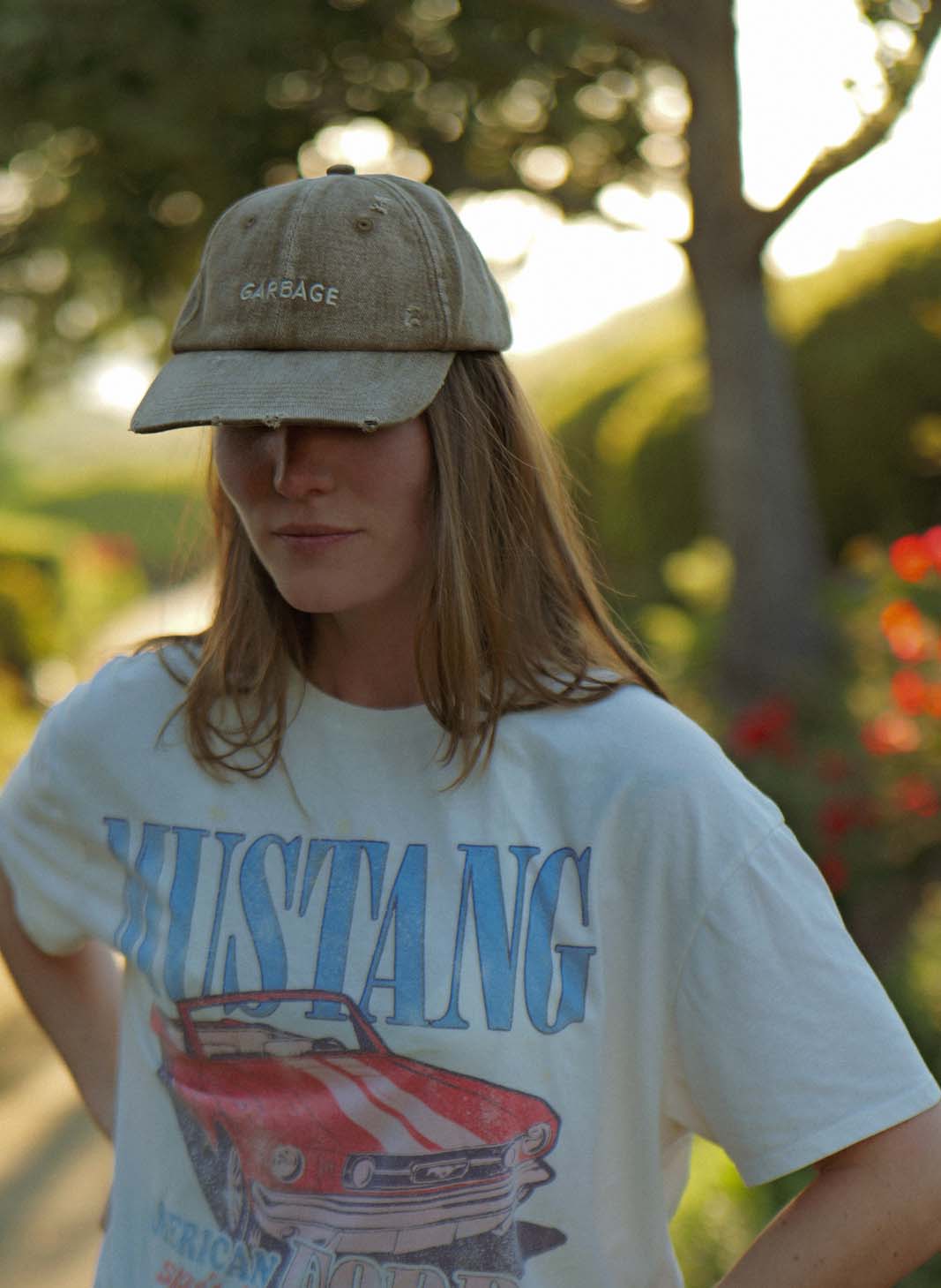 Lifestyle portrait of a person wearing the bABP B-SIDE tan washed cotton hat with "GARBAGE" embroidery and a vintage Mustang t-shirt, standing outdoors with trees in the background