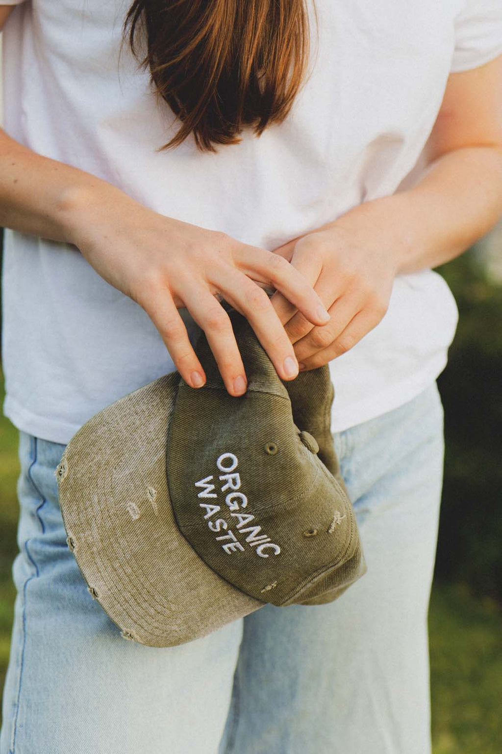Close-up lifestyle shot of a person's hands holding the bABP B-SIDE olive washed cotton hat with white "ORGANIC WASTE" embroidery, showcasing the distressed texture