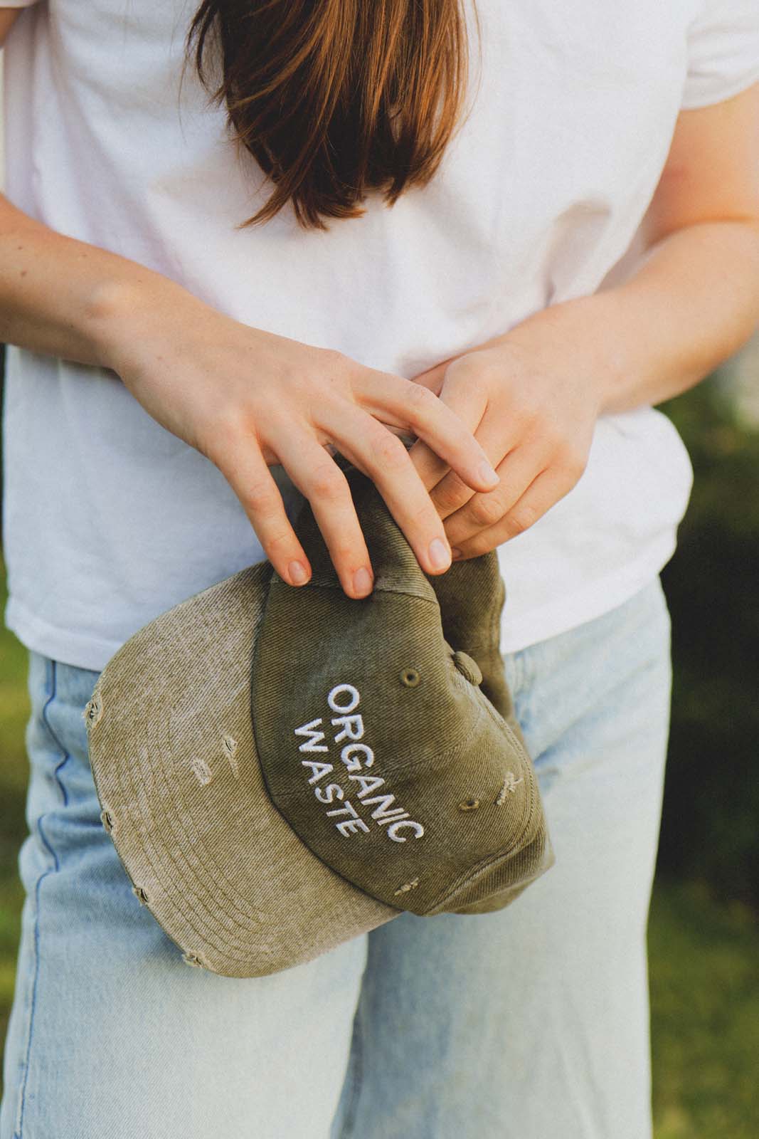Close-up lifestyle shot of a person's hands holding the bABP B-SIDE olive washed cotton hat with white "ORGANIC WASTE" embroidery, showcasing the distressed texture