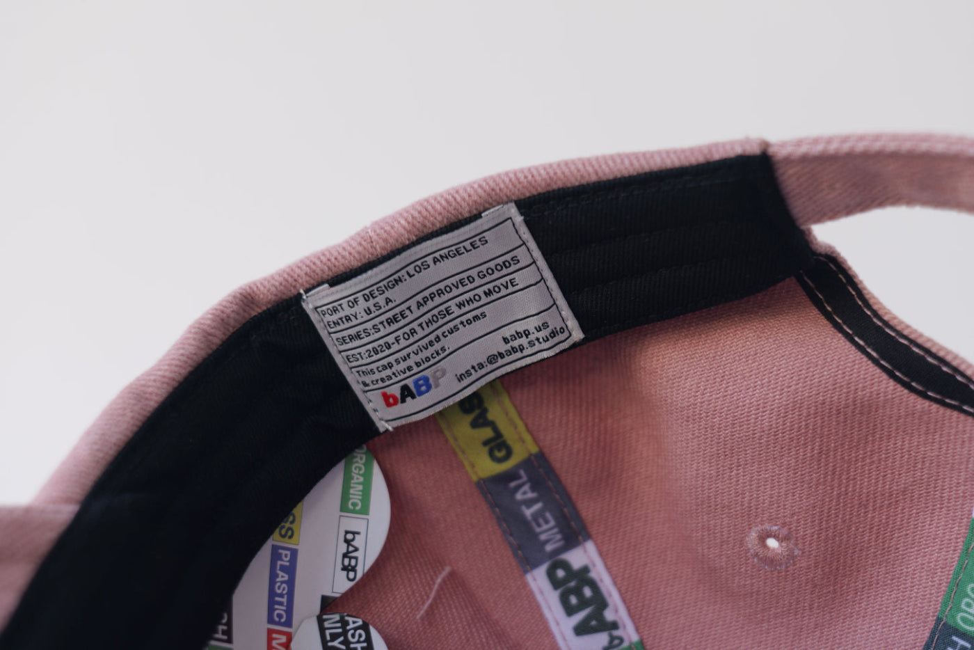 Close-up interior of the bABP B-SIDE pink heavy cotton hat showing a hidden heritage label with "Port of Design: Los Angeles" and "Street Approved Goods" text, alongside branded recycling-themed seam tapes