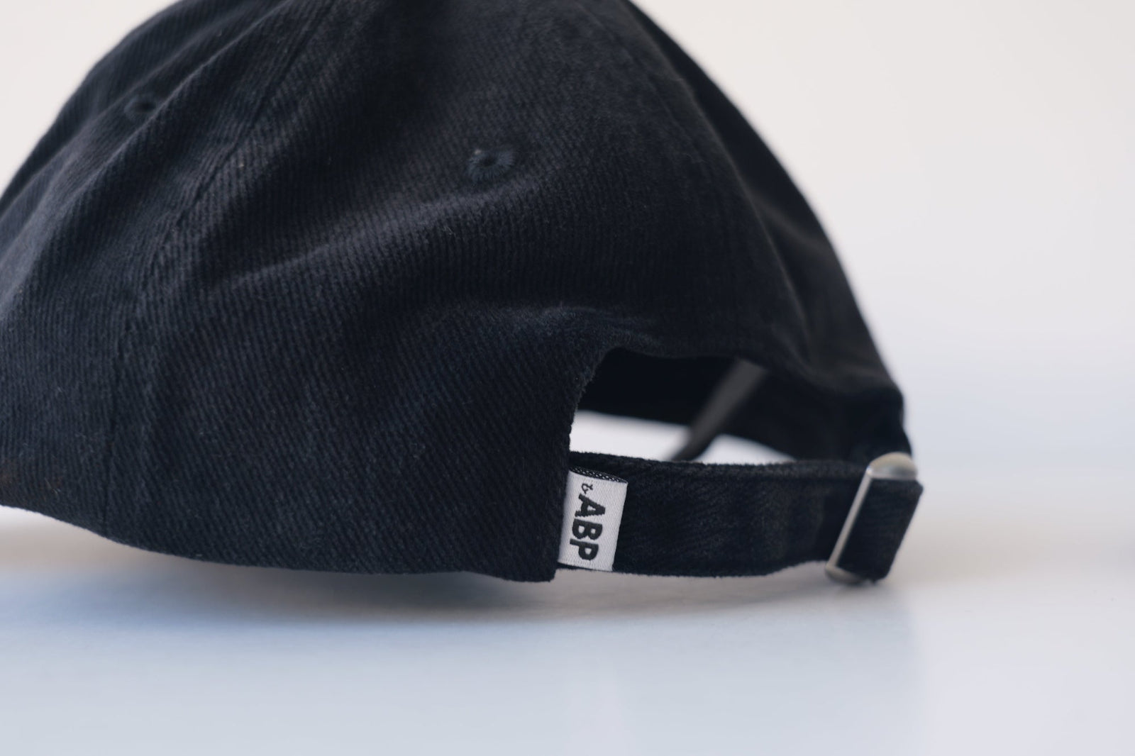 Close-up of the bABP white woven logo tag on the back of a black washed cotton hat, featuring an adjustable strap with a metal buckle, B-SIDE collection