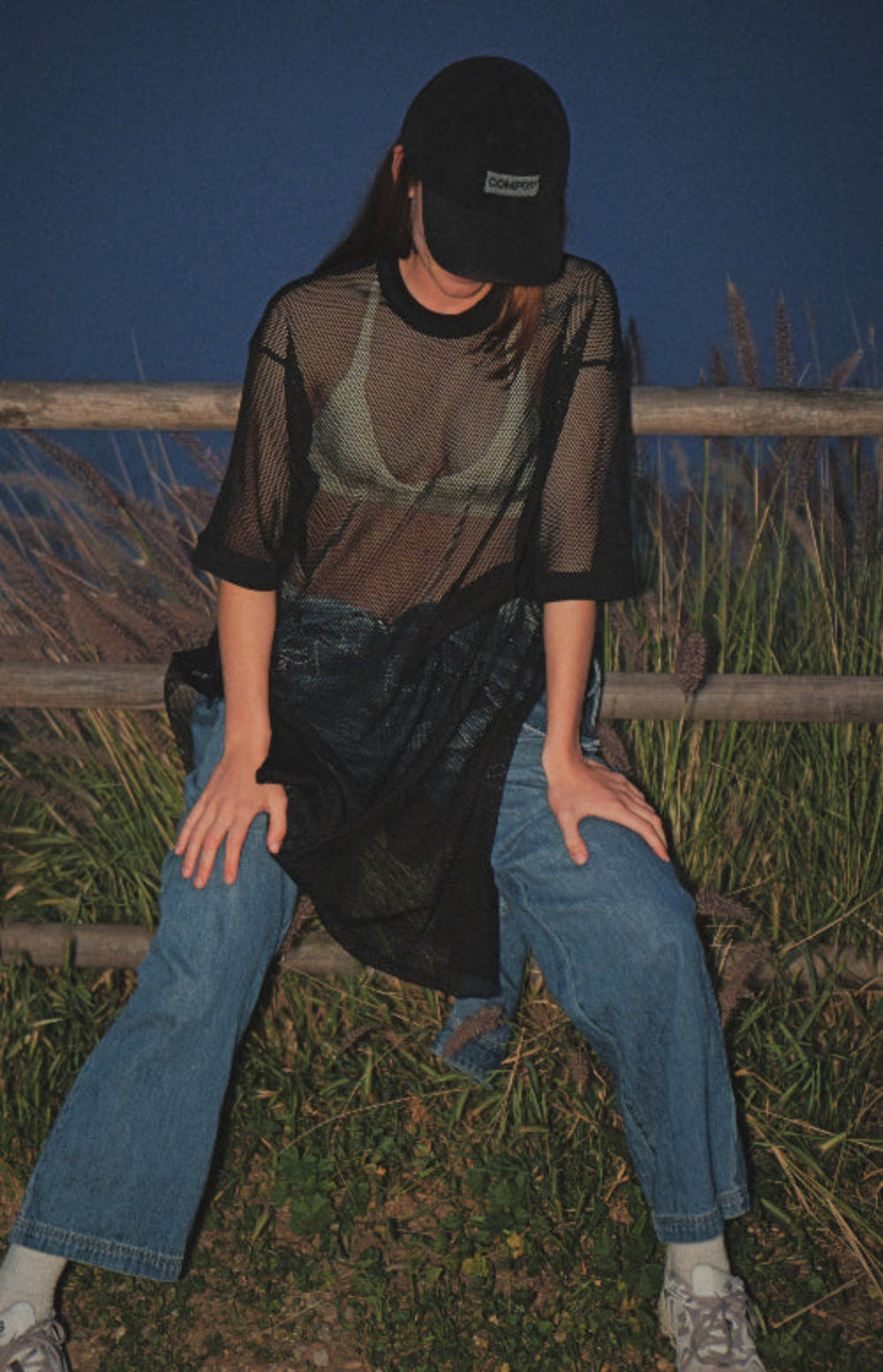 Model wearing bABP "COMPOST" black washed cotton hat with grey logo patch, styled with a sheer mesh top and jeans, leaning against a wooden fence at night, B-SIDE collection