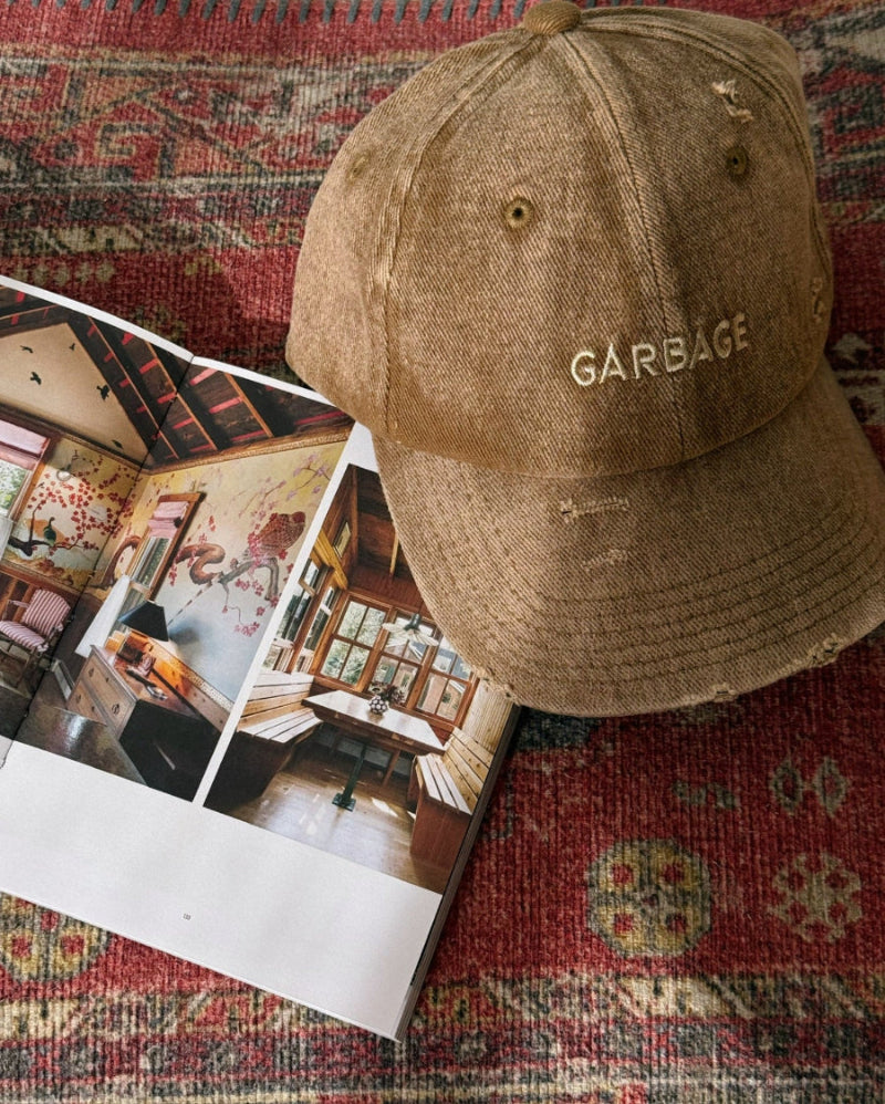 Top view of bABP "GARBAGE" tan distressed cotton dad hat with tonal beige embroidery, placed on a vintage red rug next to an open editorial magazine