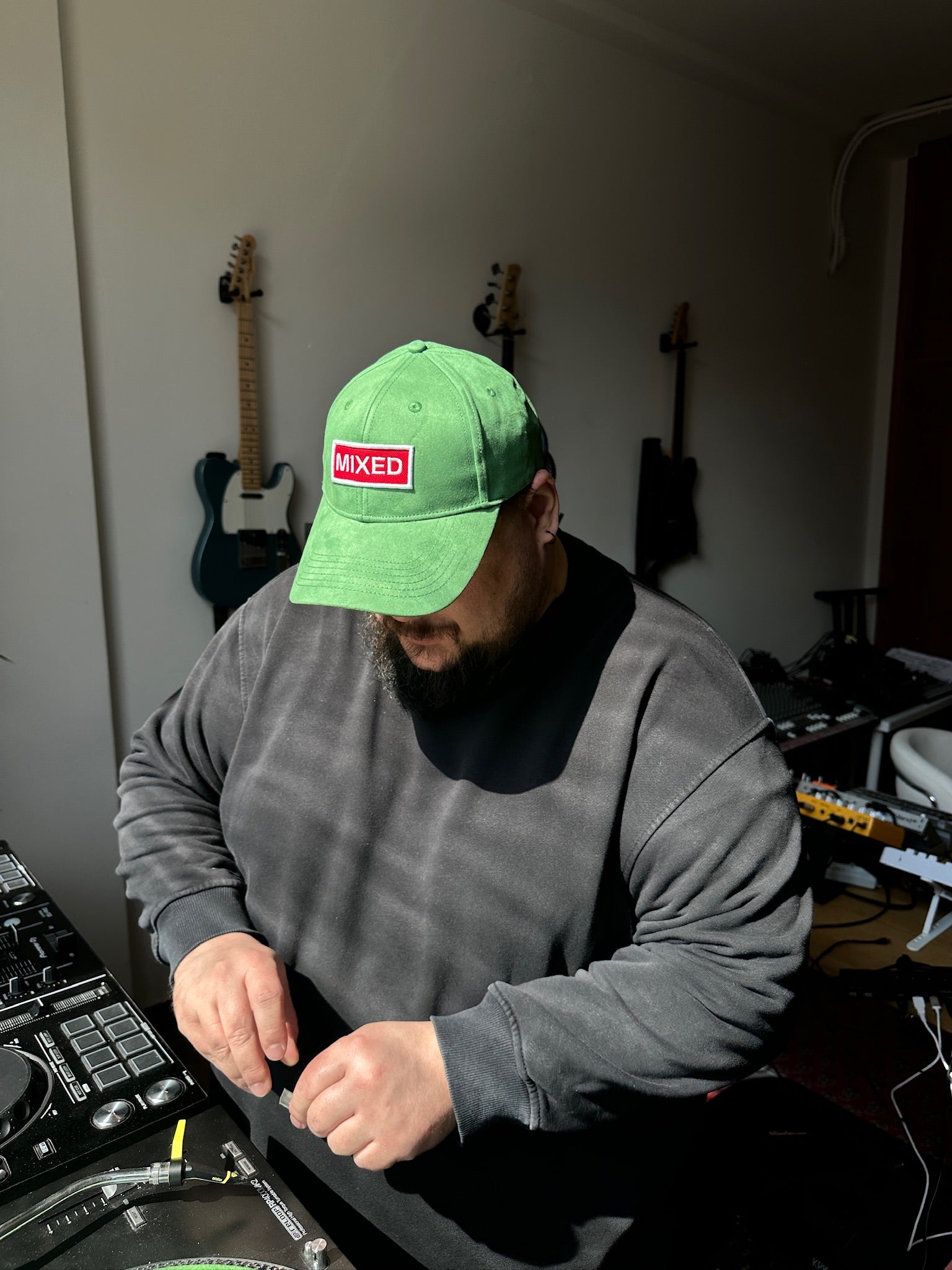 Lifestyle shot of a person wearing the bABP "MIXED" green eco-suede hat while working at a DJ mixing console in a studio with guitars in the background