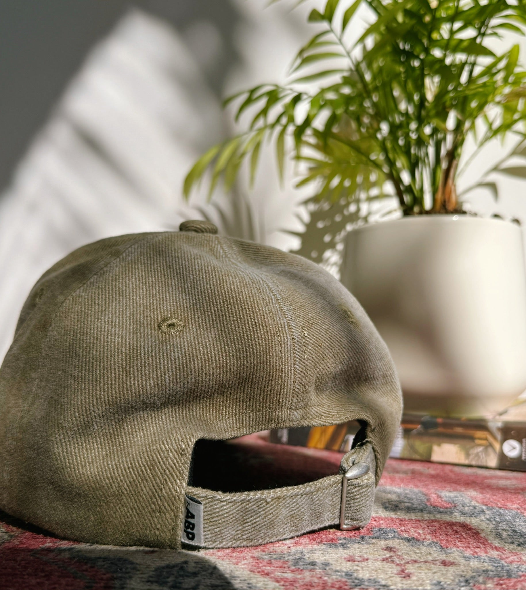Rear view of bABP "ORGANIC WASTE" sage green distressed cotton hat showing adjustable strap, metal buckle, and branded side label on a vintage rug background
