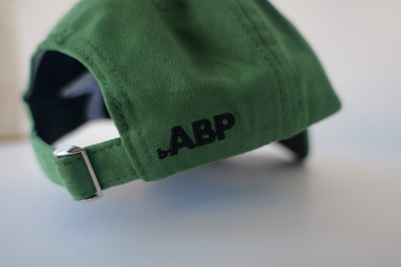 Close-up view of the back of a green eco-suede hat from the bABP Signature Collection, showing the adjustable strap with a metal buckle and a black embroidered logo on the side