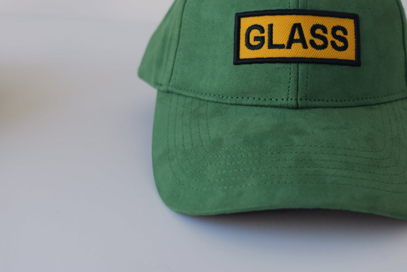Close-up front view of the bABP Signature Collection green eco-suede hat featuring a yellow embroidered patch with black "GLASS" text and premium fabric texture