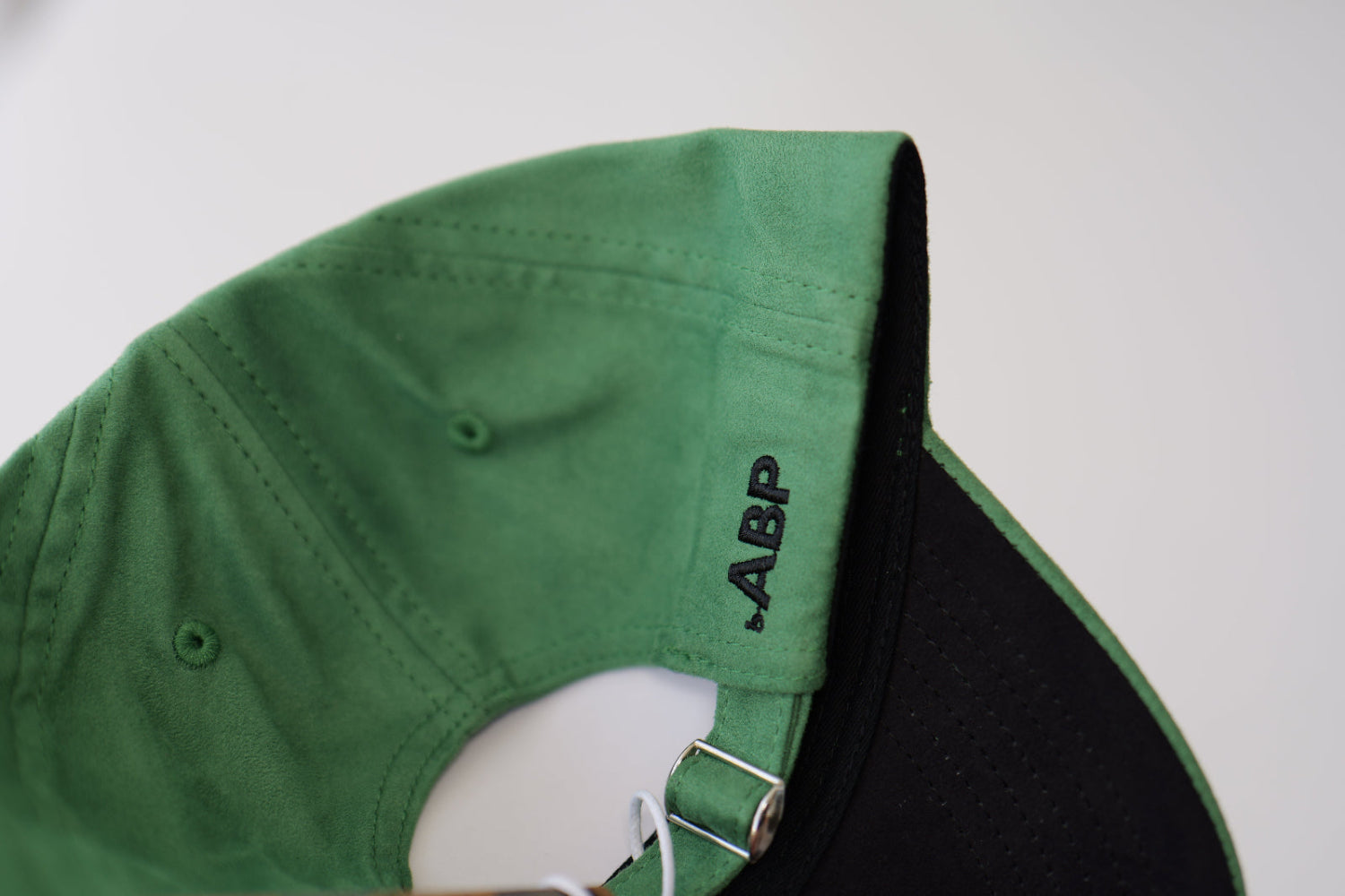 Close-up view of the back of a bABP Signature Collection green eco-suede hat, showing the black "bABP" logo embroidery on the side and the silver-tone metal adjustment buckle