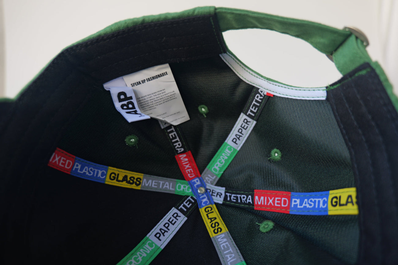 Interior view of the bABP Signature Collection green eco-suede hat showing branded "TETRA/MIXED PLASTIC/GLASS/METAL/PAPER" seam tapes, "SPEAK UP FASHIONABLE" woven label, and a metal buckle on the adjustable strap