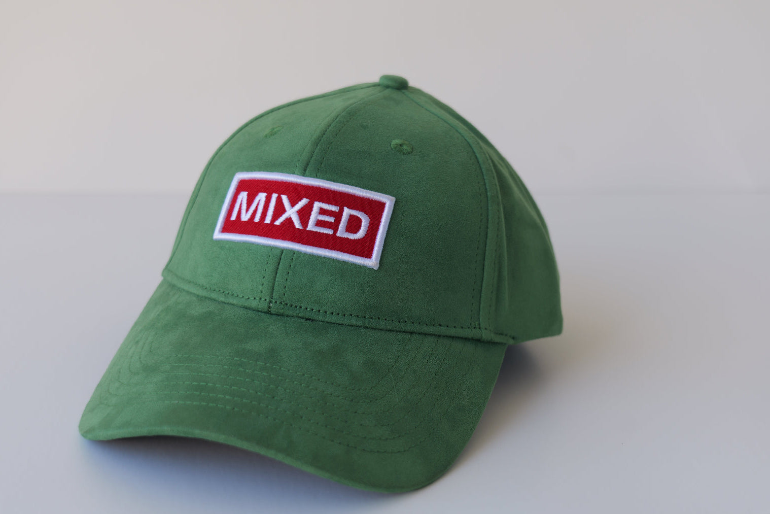 Studio shot of the bABP Signature Collection green eco-suede hat featuring a red and white "MIXED" embroidered patch on a premium textured emerald background