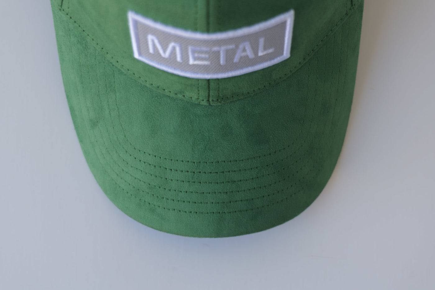 Front view of the bABP Signature Collection green eco-suede hat featuring a light gray embroidered patch with white "METAL" text and premium fabric texture