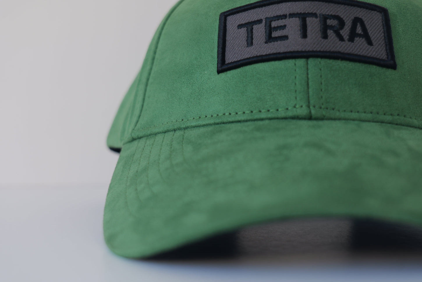 Extreme close-up of the bABP Signature Collection green eco-suede hat showing a dark gray embroidered patch with a black "TETRA" logo and black border