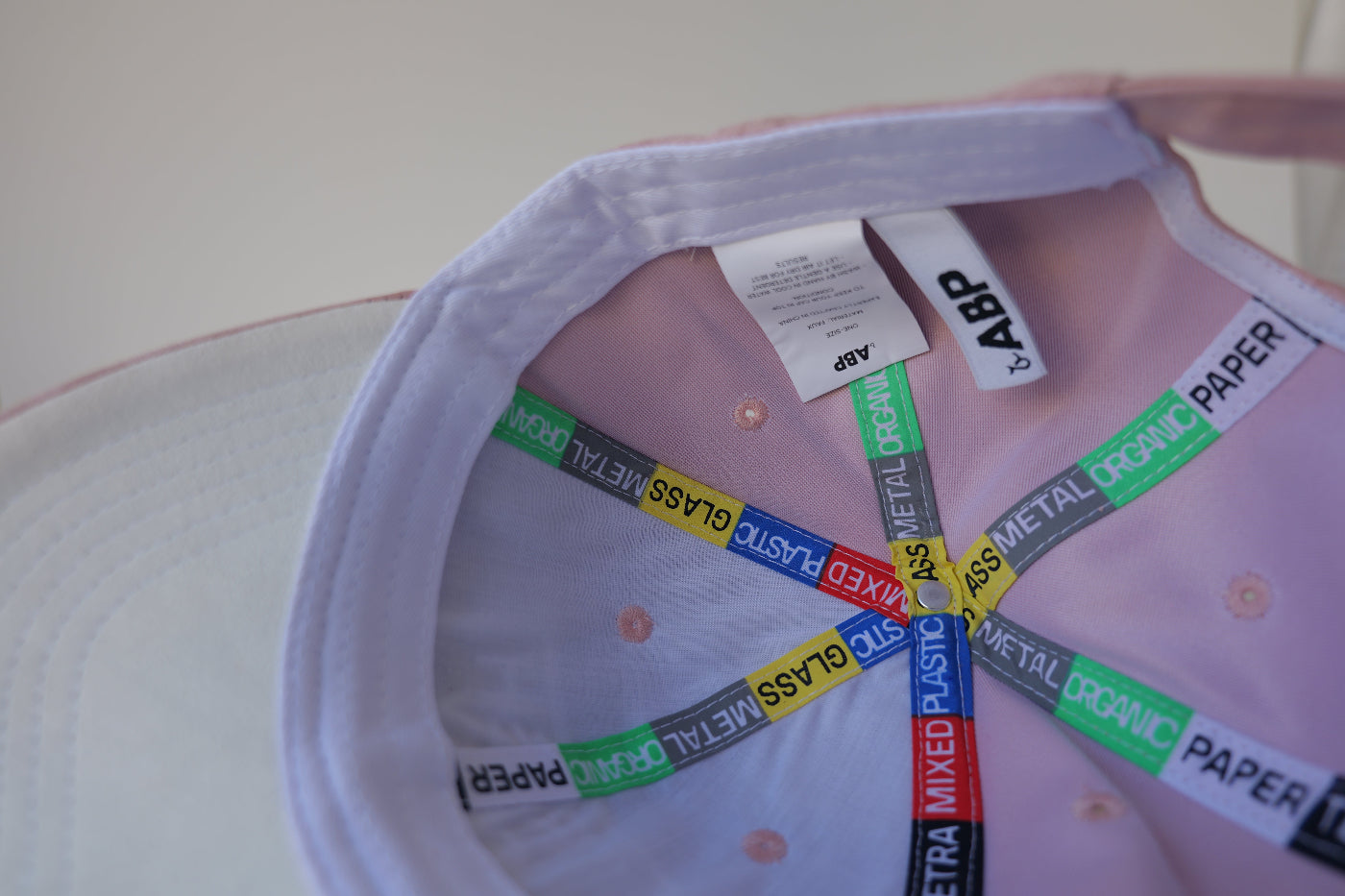 Interior close-up of the bABP Signature Collection pink eco-suede hat featuring two branded woven labels, "TRASH ONLY" identity, and "PLASTIC METAL GLASS" recycling-themed seam tapes