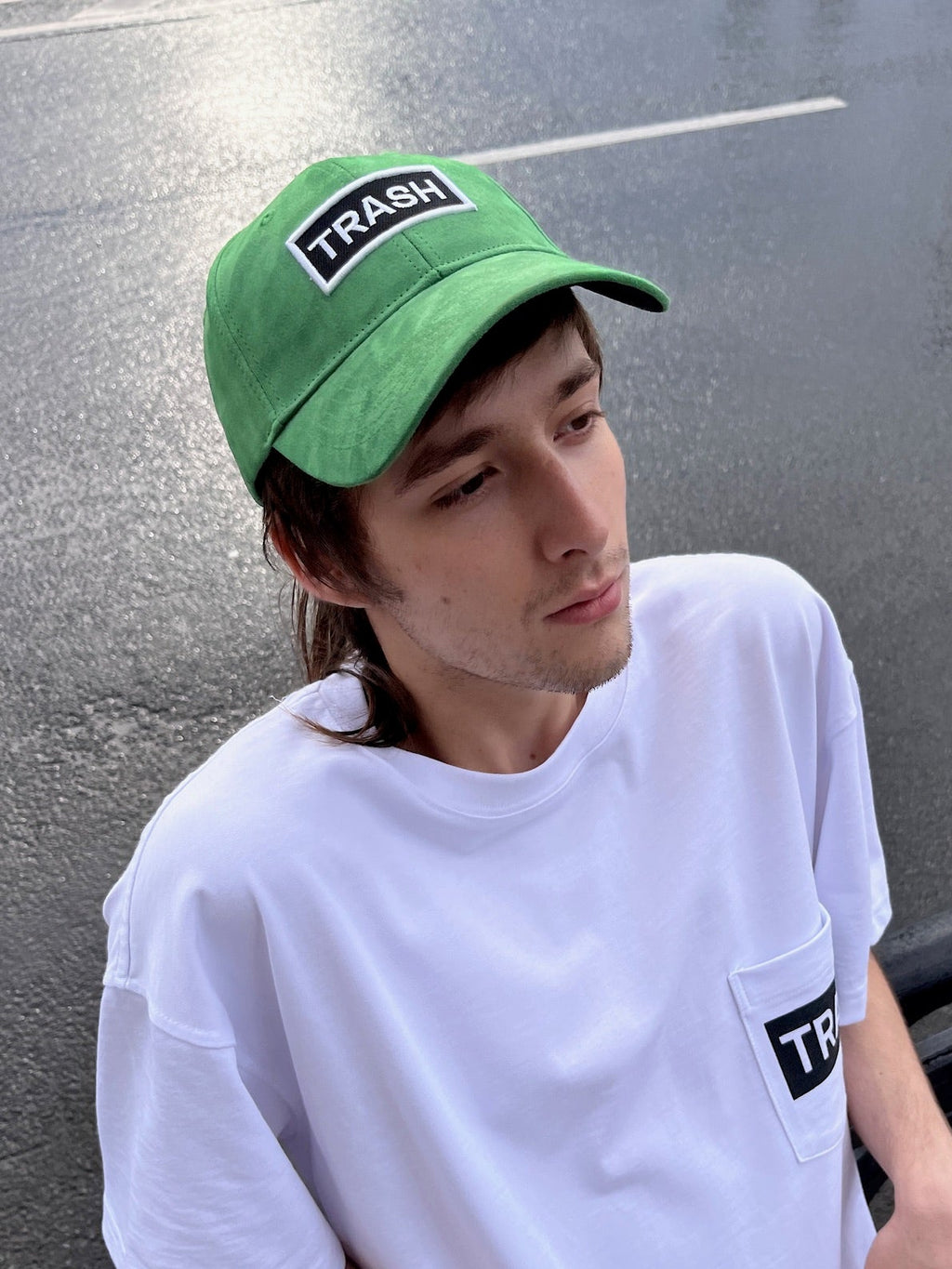 Lifestyle shot of a person wearing the bABP "TRASH" green eco-suede signature hat and a white pocket t-shirt, leaning against a black metal fence on a city street